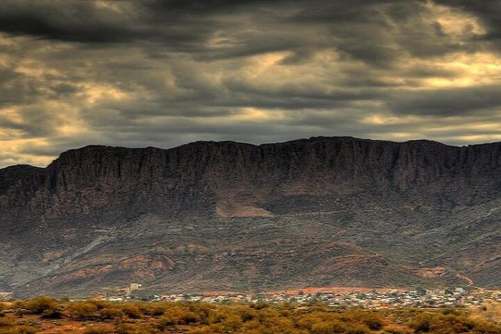 A dramatic sky with dark clouds looms over a rugged mountain range, with a small town and sparse vegetation in the foreground.