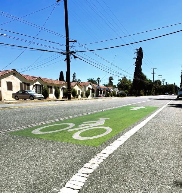 A green bike lane is marked on a residential street, with houses, power lines, and trees visible under a clear blue sky.