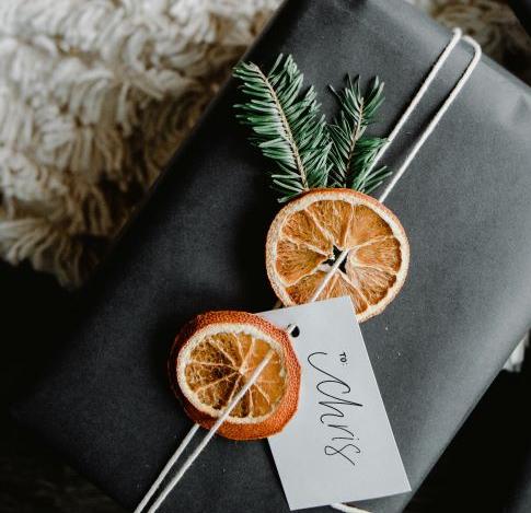 A black gift box tied with white string, decorated with dried orange slices, pine sprigs, and a gift tag labeled “Chris.”.