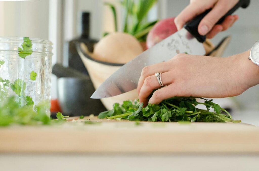A person chops fresh cilantro on a wooden cutting board with a knife, next to a glass jar and a bowl of vegetables in the background.
