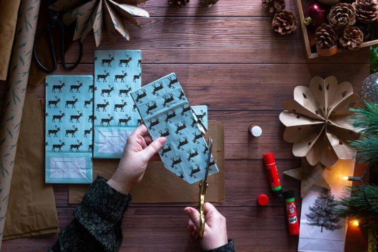 Person cutting blue paper with reindeer pattern on a wooden table, surrounded by craft supplies, glue sticks, pinecones, and holiday decorations.