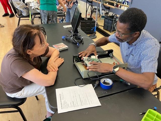 A man repairs an electronic device while a woman observes at a table covered with tools, paperwork, and tape in a workshop setting.
