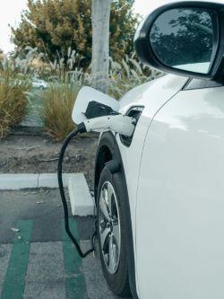 A white electric car is parked and charging at a charging station, with the charger plugged into the vehicle's side.