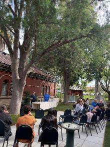 People are seated outdoors listening to two speakers standing at a table in front of a brick building, surrounded by trees.