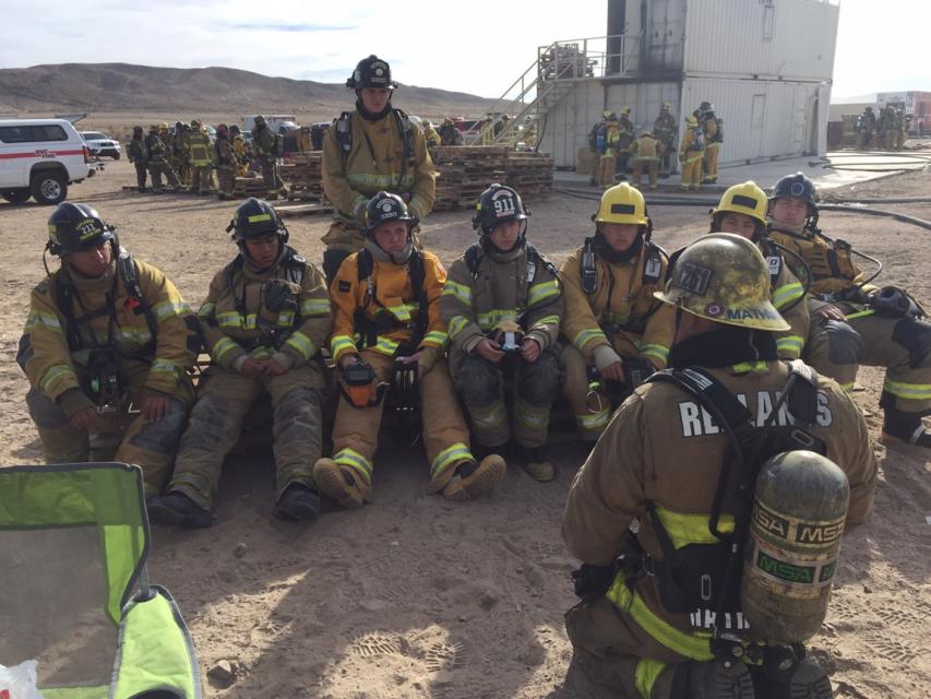 A group of firefighters in full gear sit on the ground while another firefighter stands facing them, with training buildings and equipment visible in the background.