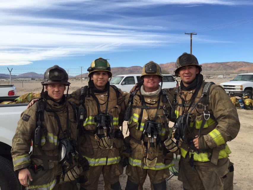 Four firefighters in full gear stand together outdoors in front of utility vehicles and desert landscape, posing for a group photo.