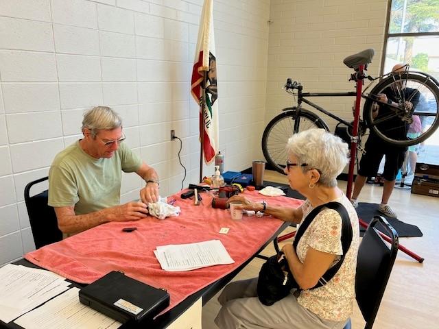 An older man repairs a small object at a table covered with papers and tools while an older woman watches; a bicycle is being serviced in the background.