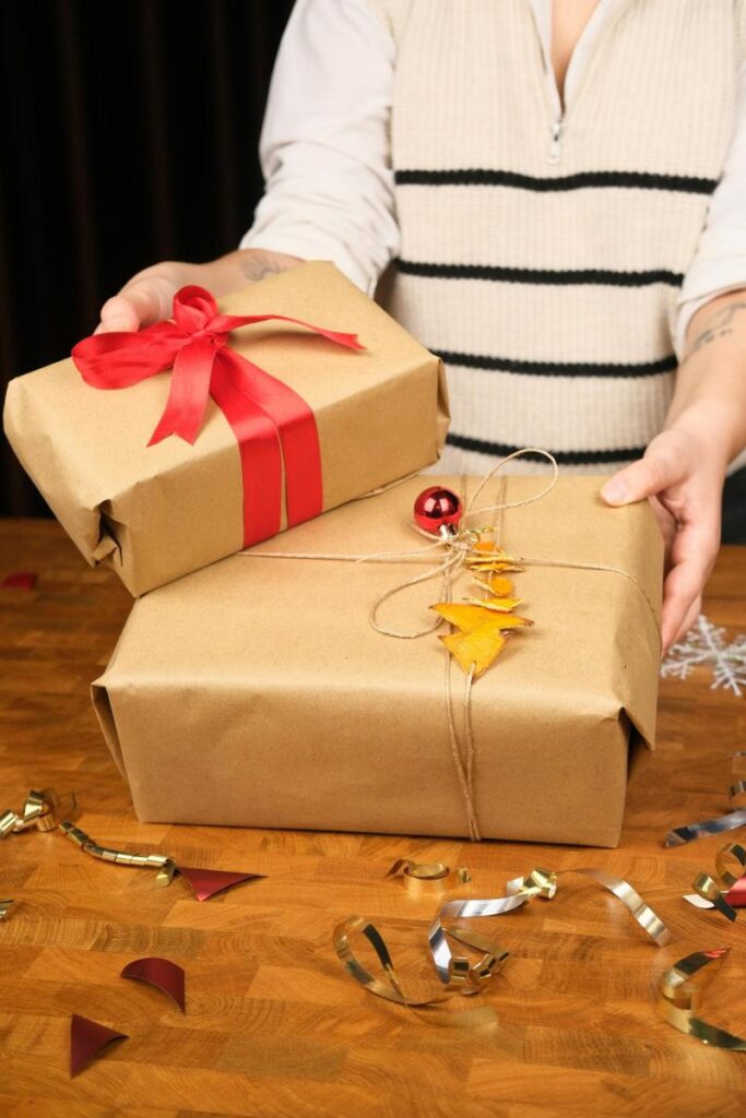 Person holding two brown paper-wrapped gifts, one with a red ribbon and bow, the other with string, yellow leaves, and a red ornament, on a wooden table with festive decorations.