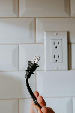 A hand holding a black electrical plug in front of a white wall outlet with a tiled backsplash.