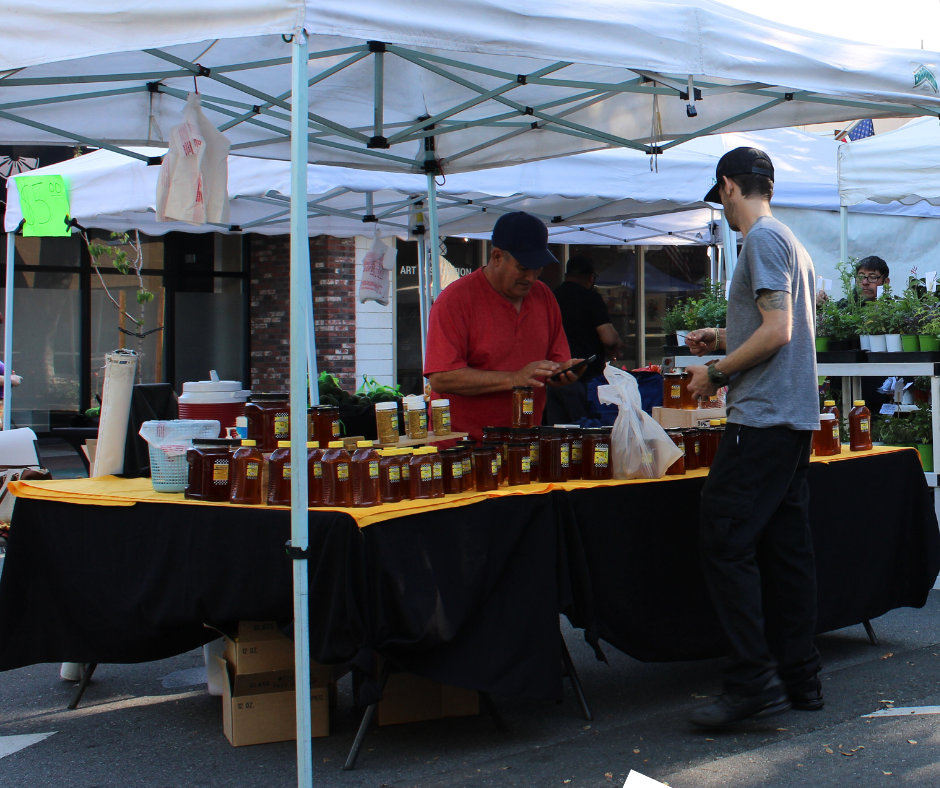 Two men stand under a market tent arranging and purchasing jars of honey displayed on tables, with plants for sale in the background.