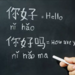 Hand holding chalk near a blackboard shows Chinese phrases with pinyin: “nǐ hǎo” equals “Hello” and “nǐ hǎo ma” equals “How are you.”.