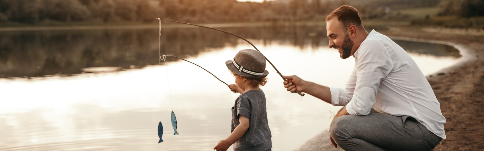An adult and a child wearing a hat are fishing together by a calm lakeshore, holding fishing rods with toy fish attached.