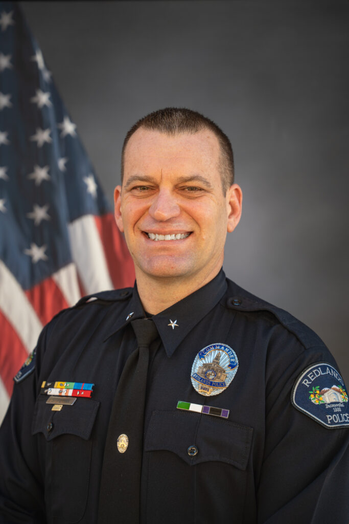 A police officer in formal uniform stands in front of an American flag, smiling at the camera.