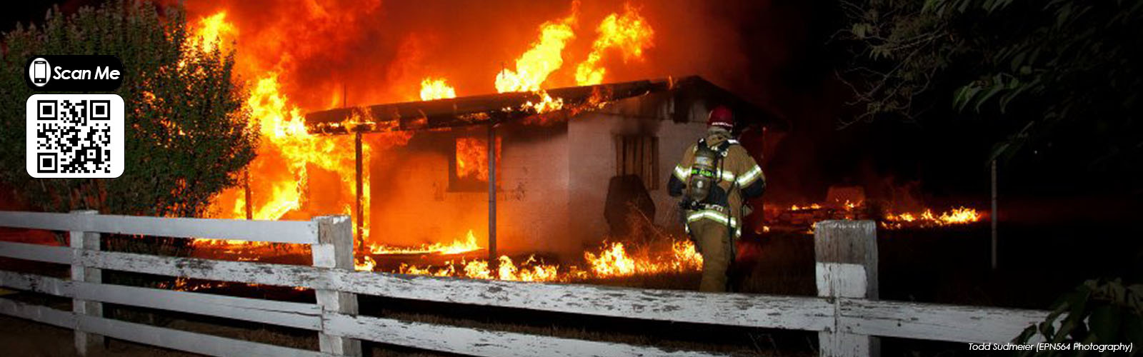 A firefighter approaches a burning house at night, with flames engulfing the roof and yard. A white wooden fence runs along the foreground.