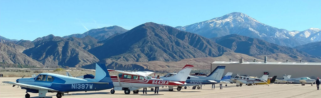 Small aircraft are parked in a row on an airport tarmac with people standing nearby, mountains and a clear sky in the background.
