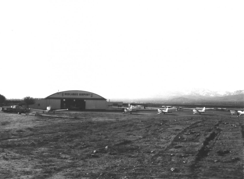 A small airfield with a hangar labeled "Redlands Airport" and several small airplanes parked on a dirt field, mountains visible in the background.