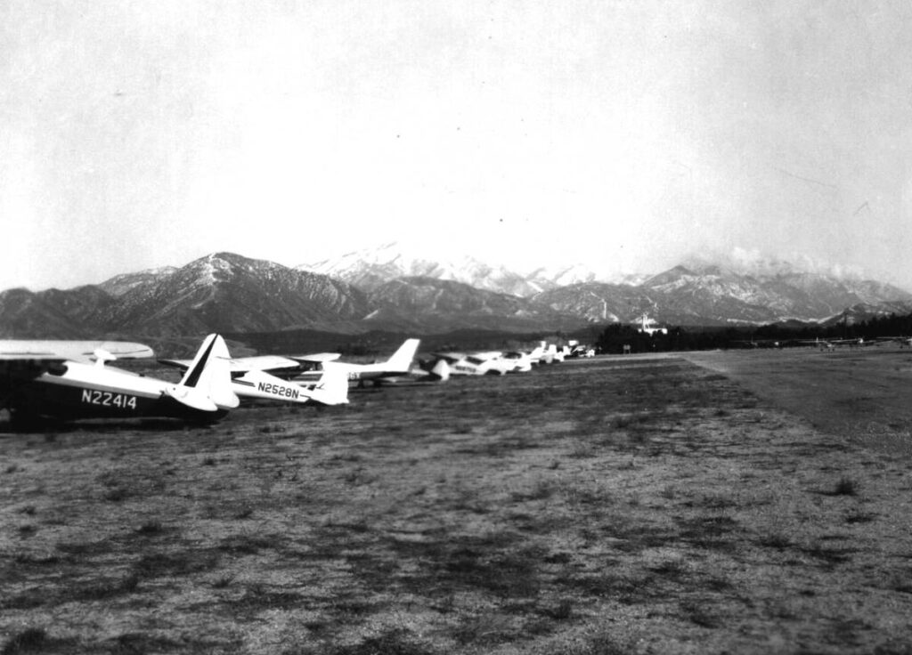 Several small airplanes are parked in a row on a grassy airfield with snow-capped mountains visible in the background.