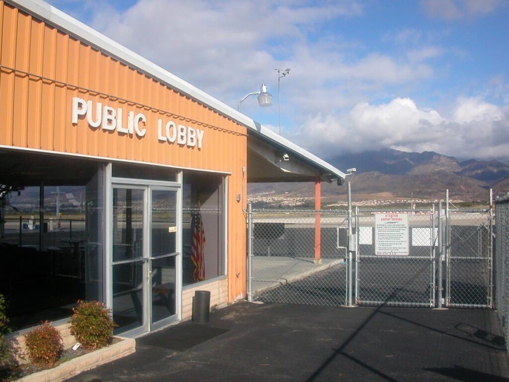 Orange public lobby building with glass doors next to a fenced gate and sign, mountains and cloudy sky in the background.