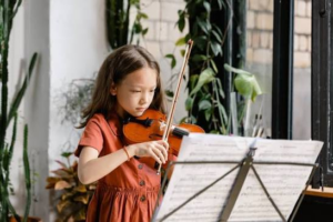 A young girl in a red dress plays the violin indoors, reading sheet music from a stand, with plants and large windows in the background.