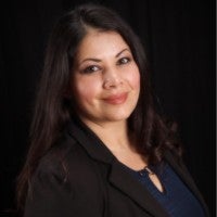 Woman with long dark hair wearing a black blazer and blue blouse, smiling in front of a dark background.